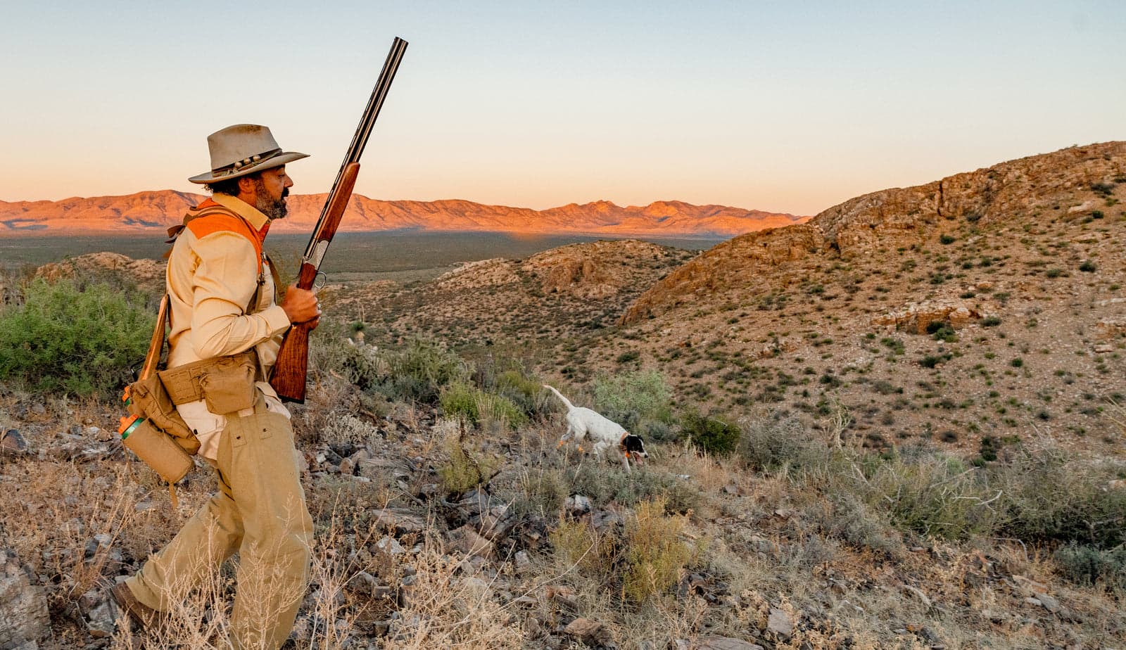 Man wearing a hat and beige outdoor clothing holding a rifle and walking through a desert landscape at sunset with a hunting dog nearby.