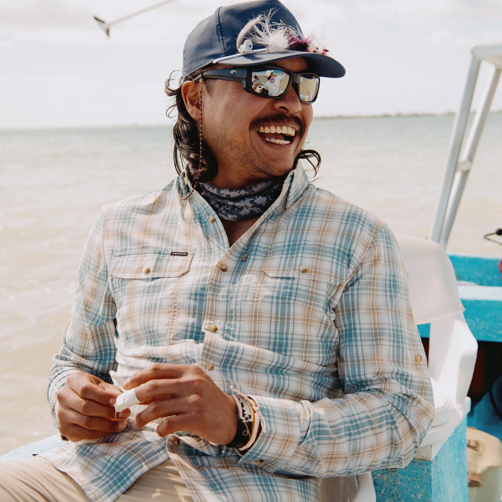 Man wearing a light plaid shirt, sunglasses, and a cap with fishing flies, smiling on a boat near water.